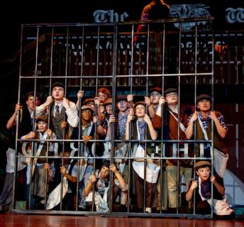 Newsies cast behind tall “The World” stage gate during performance.